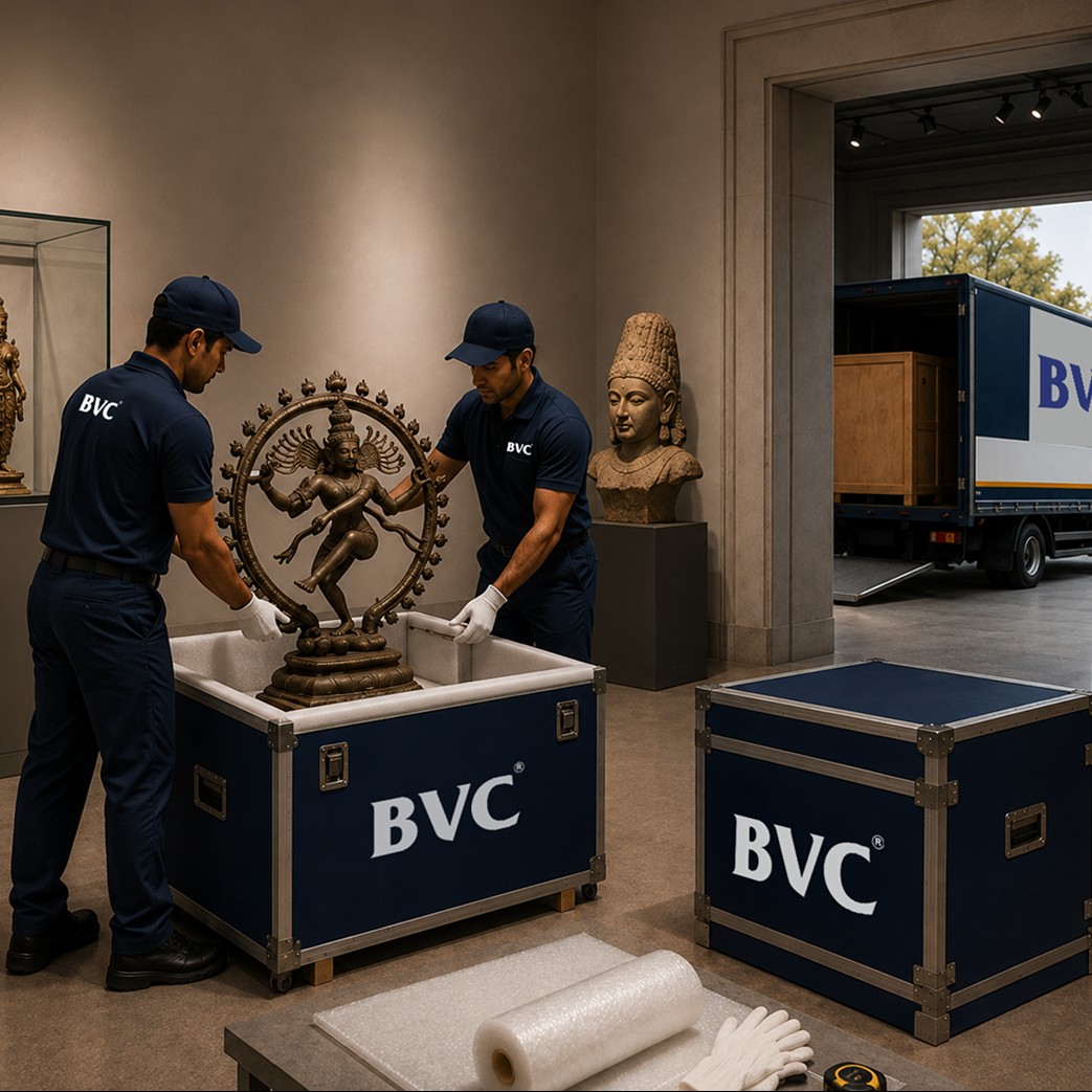 Two BVC handlers in navy uniforms and white gloves crating a bronze Nataraja sculpture into a foam-lined blue BVC art crate in a museum gallery, with a glass vitrine, a stone bust, packing materials and a blue BVC art truck backed up at the loading door